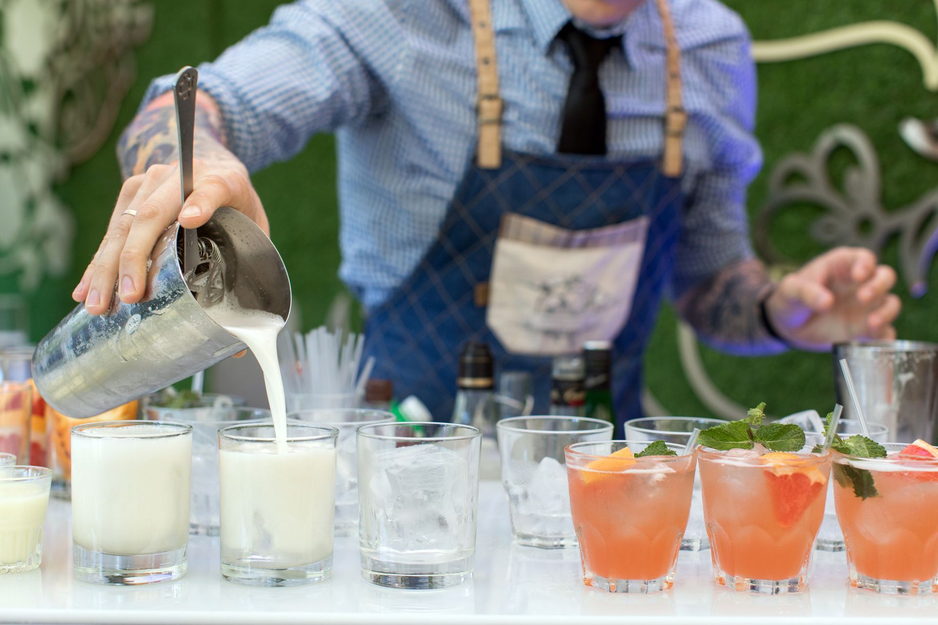 A bartender wearing a blue apron pours a white cocktail from a metal shaker into a glass, next to rows of colorful drinks.