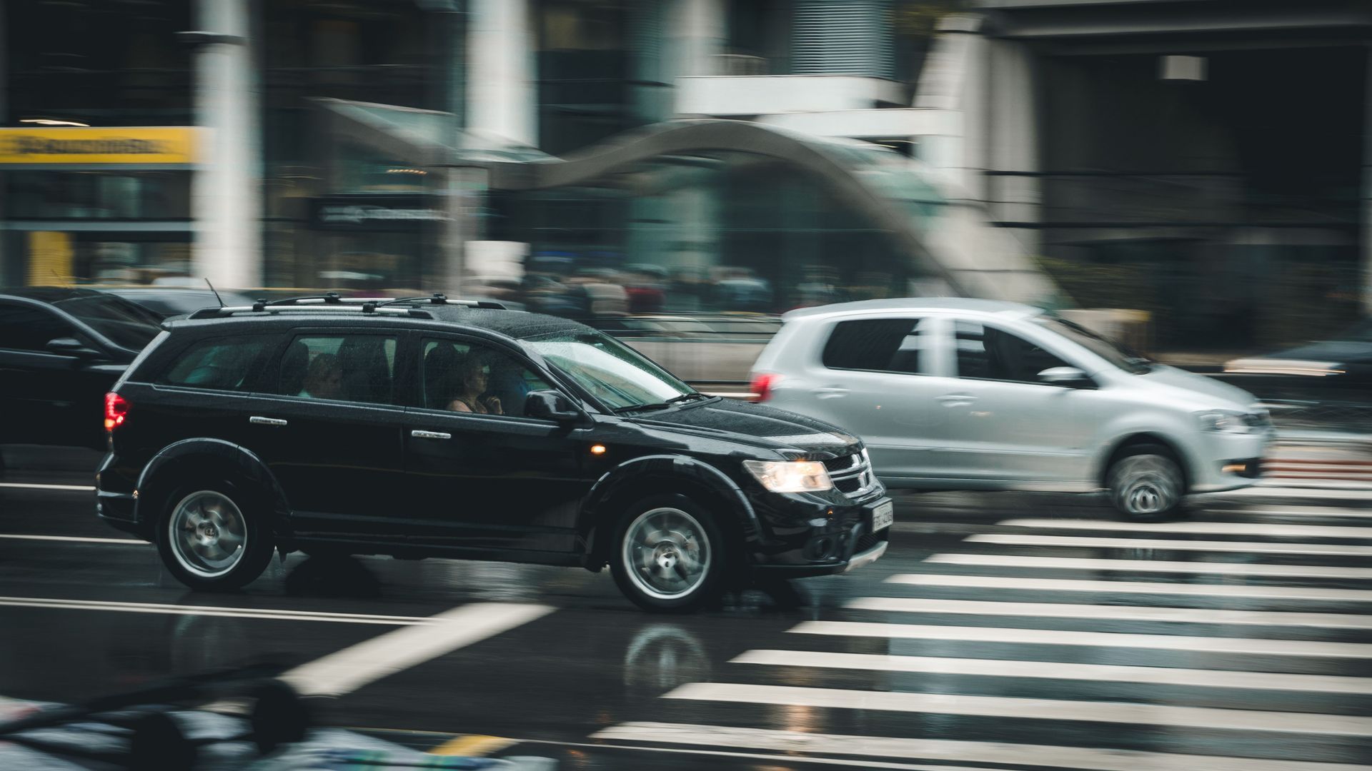 Black SUV and silver car driving across a wet crosswalk in a city intersection