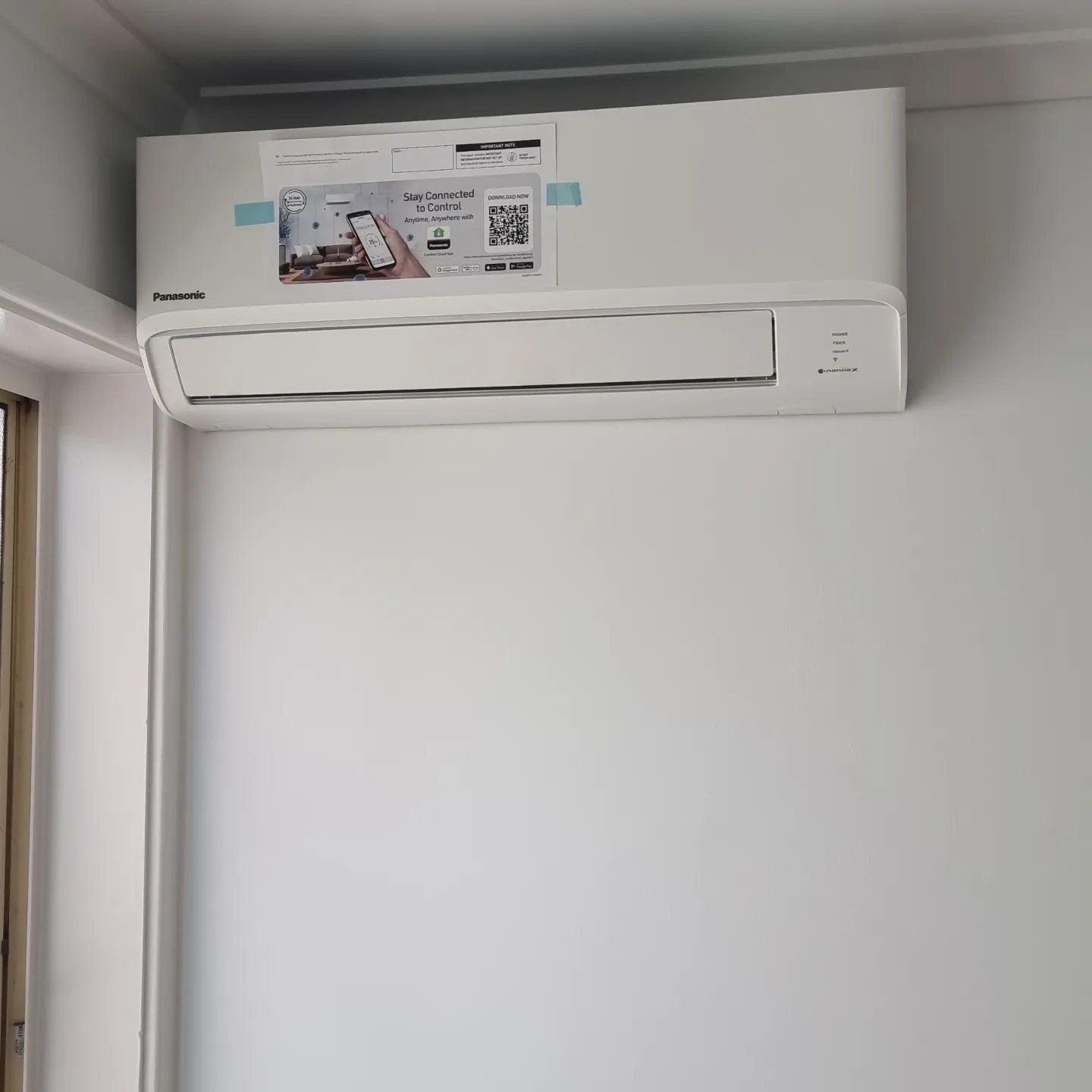A White Air Conditioner Is Mounted On A White Wall Next To A Window — Rapidcool Air Conditioning & Electrical In Dapto, NSW