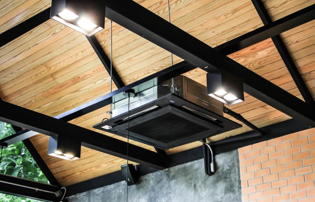 A cassette-style air conditioning unit mounted to the ceiling with timber trusses and lighting.