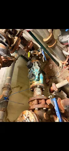 Rusty industrial pipes and valves in a cramped mechanical room, with a corroded central valve assembly.