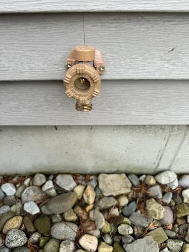 Brass outdoor water spigot mounted on gray house siding above a stone border