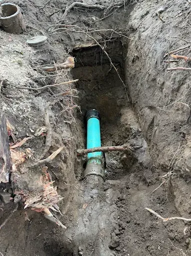 Trench exposing a blue underground pipe in muddy soil, with roots and a small opening above it