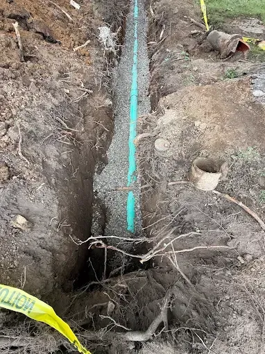 Trench with turquoise pipe and gravel lining beside a large hole in muddy ground