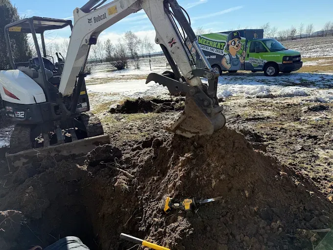 Excavator digging a muddy trench in a snowy field, with a worker in a green vehicle nearby