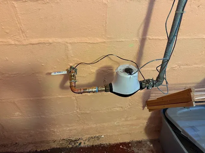 Exposed wall plumbing with a wrapped pipe, wires, and a white vent opening beside a bed
