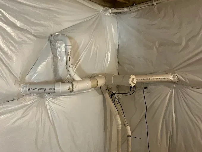 Plastic-covered crawlspace with exposed pipes, fittings, and a vent duct on the wall.