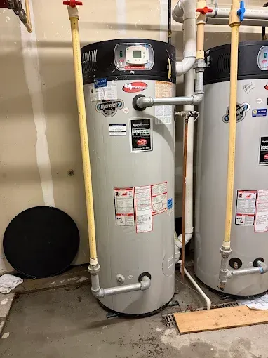 Two gray water heaters with pipes and valves in a utility room, one with a black vent cover behind it
