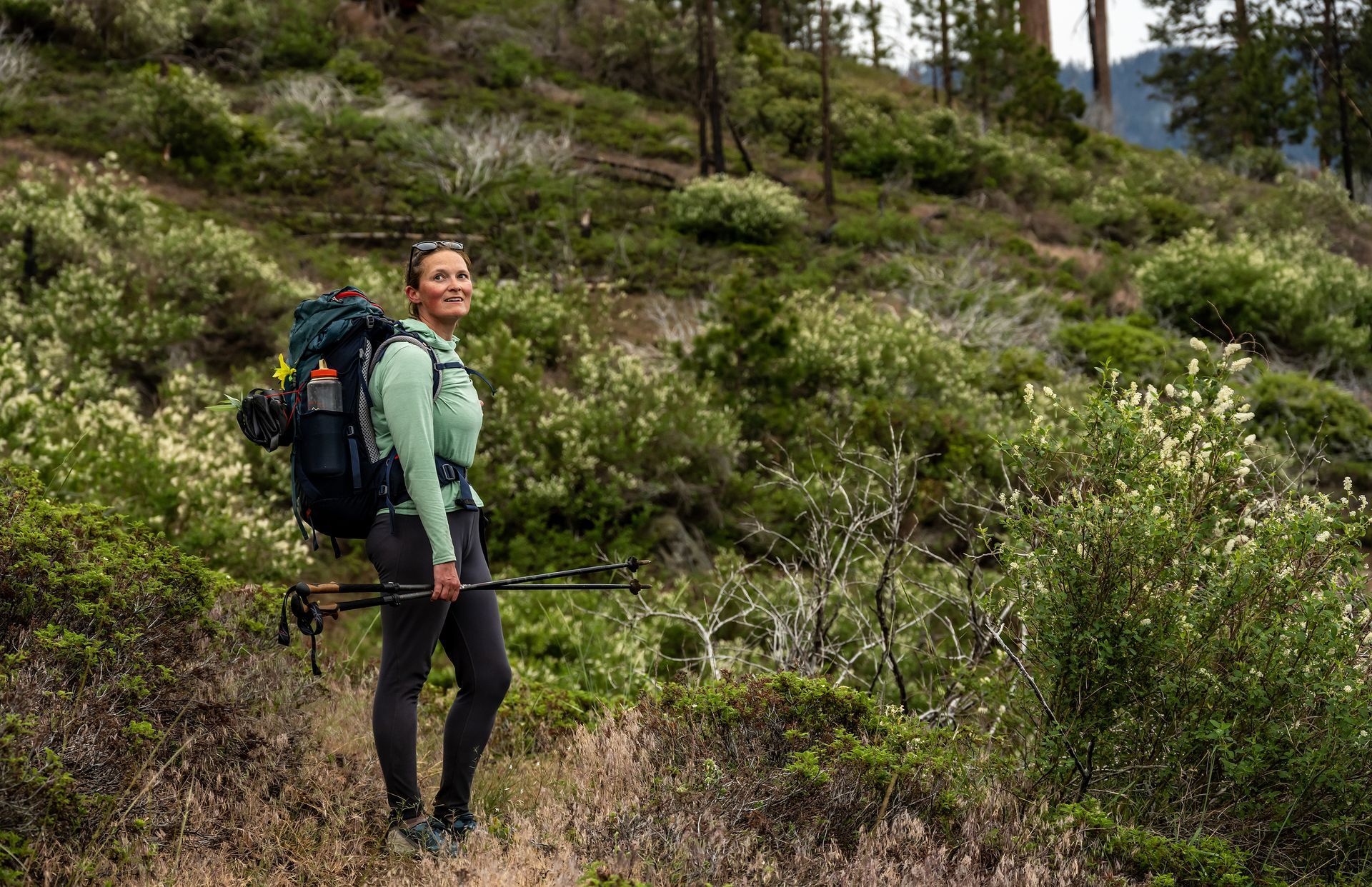 A woman with a backpack and hiking poles is standing in the woods.