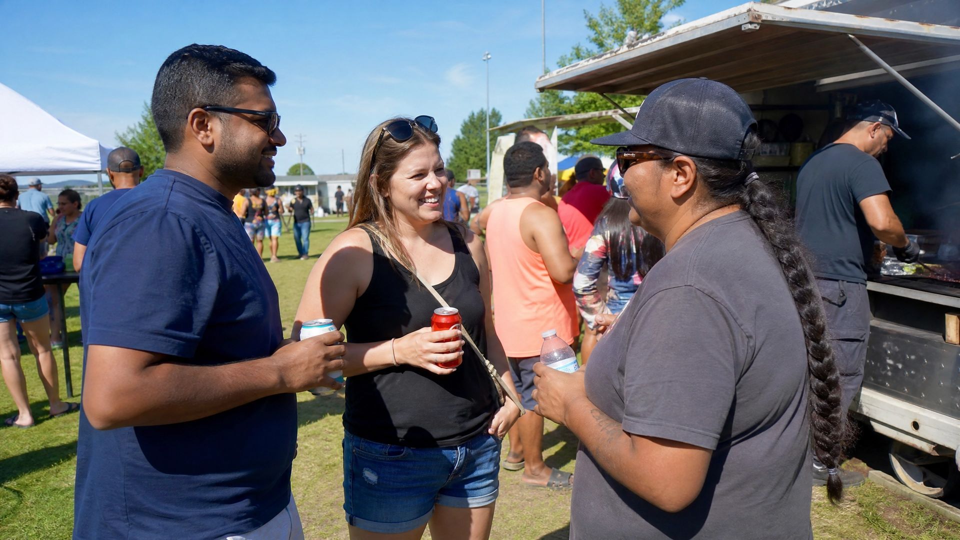 Three people chatting at an outdoor gathering near a pavilion, holding drinks in the sun.