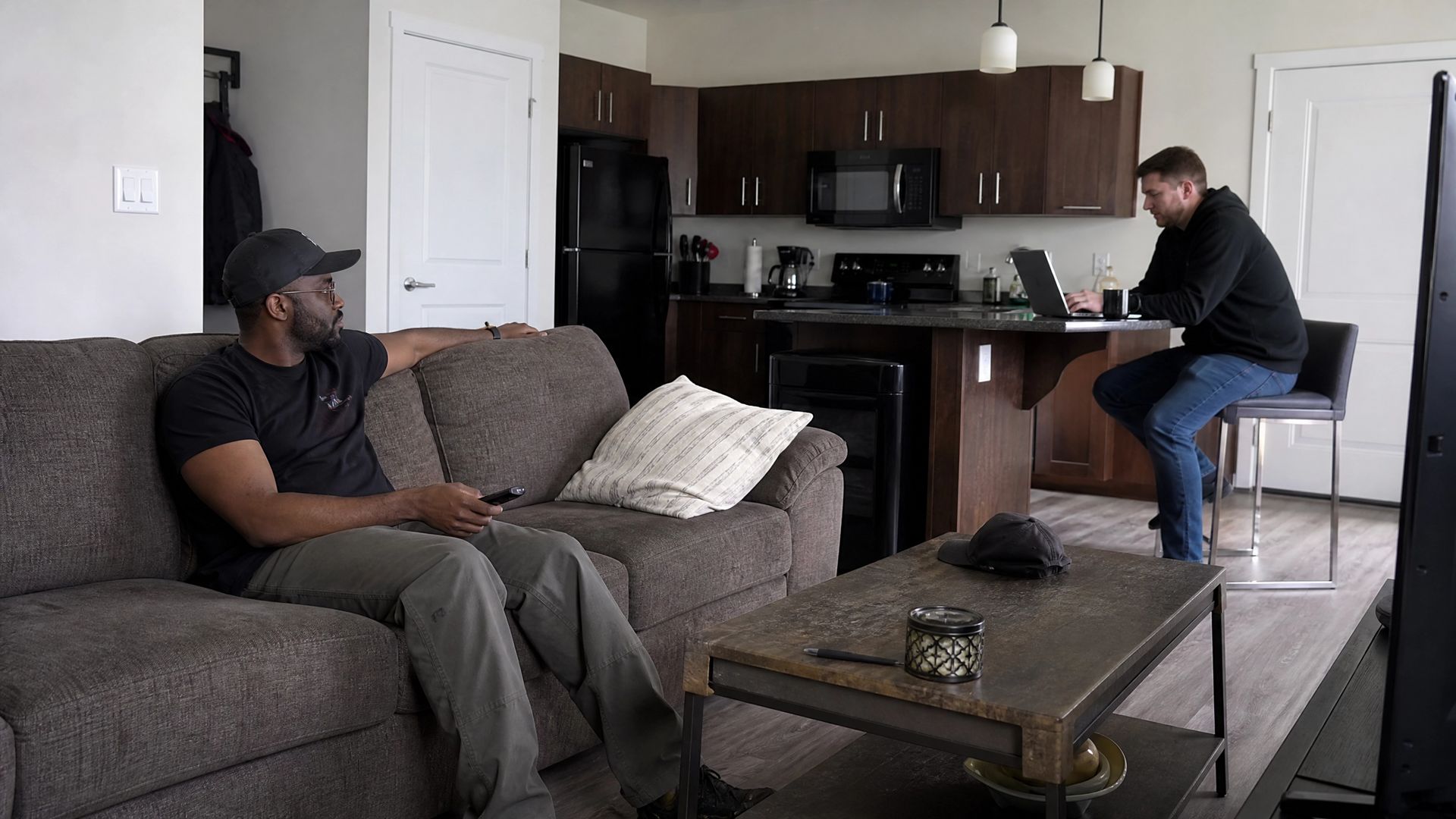 Two people relaxing in a living room with an open kitchen, one on a couch and one seated at the island.