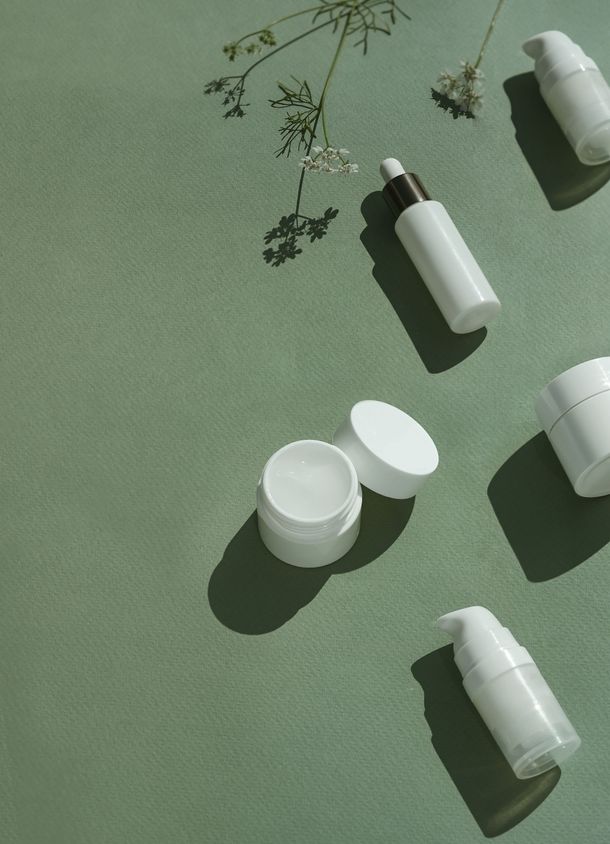 White cosmetic jars and tubes arranged on a green background with a sprig of flowers.