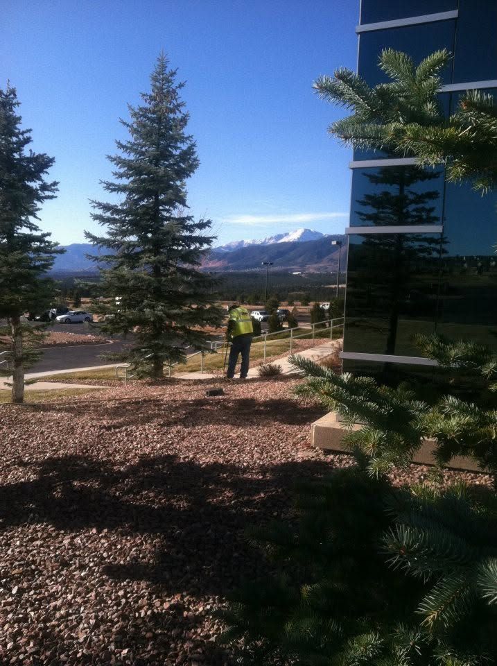 Mountain view through evergreens, with two people standing in a landscaped outdoor area beside a building.