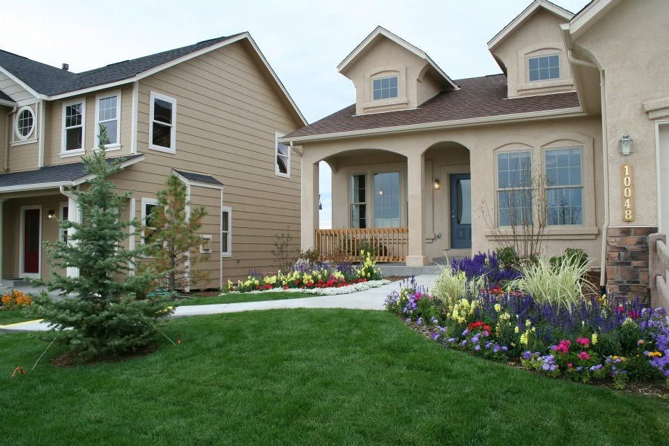 Suburban homes with manicured lawns and colorful flower gardens under a partly cloudy sky