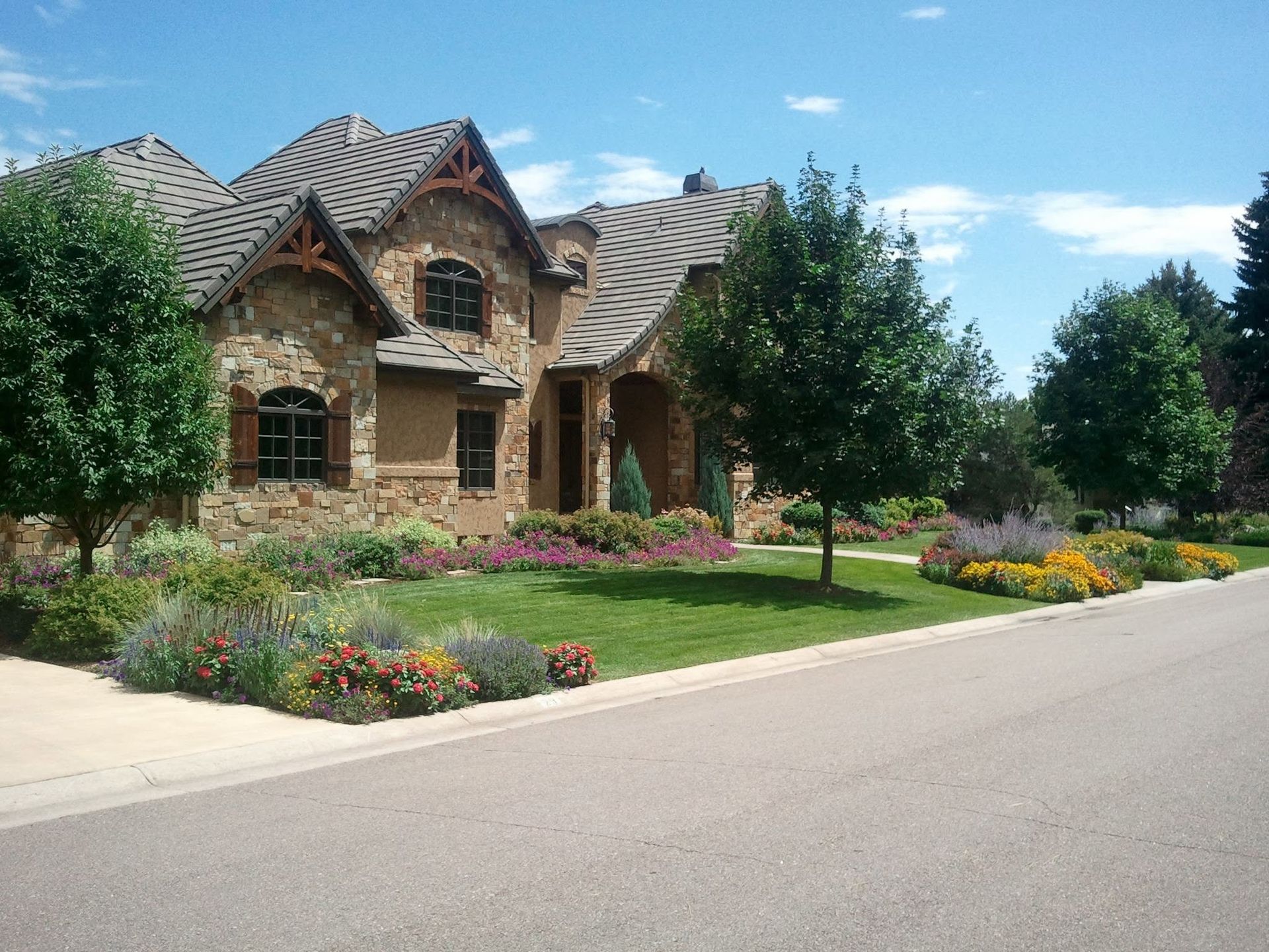 Stone house with a gray roof, manicured lawn, colorful flower beds, and trees along a quiet street.