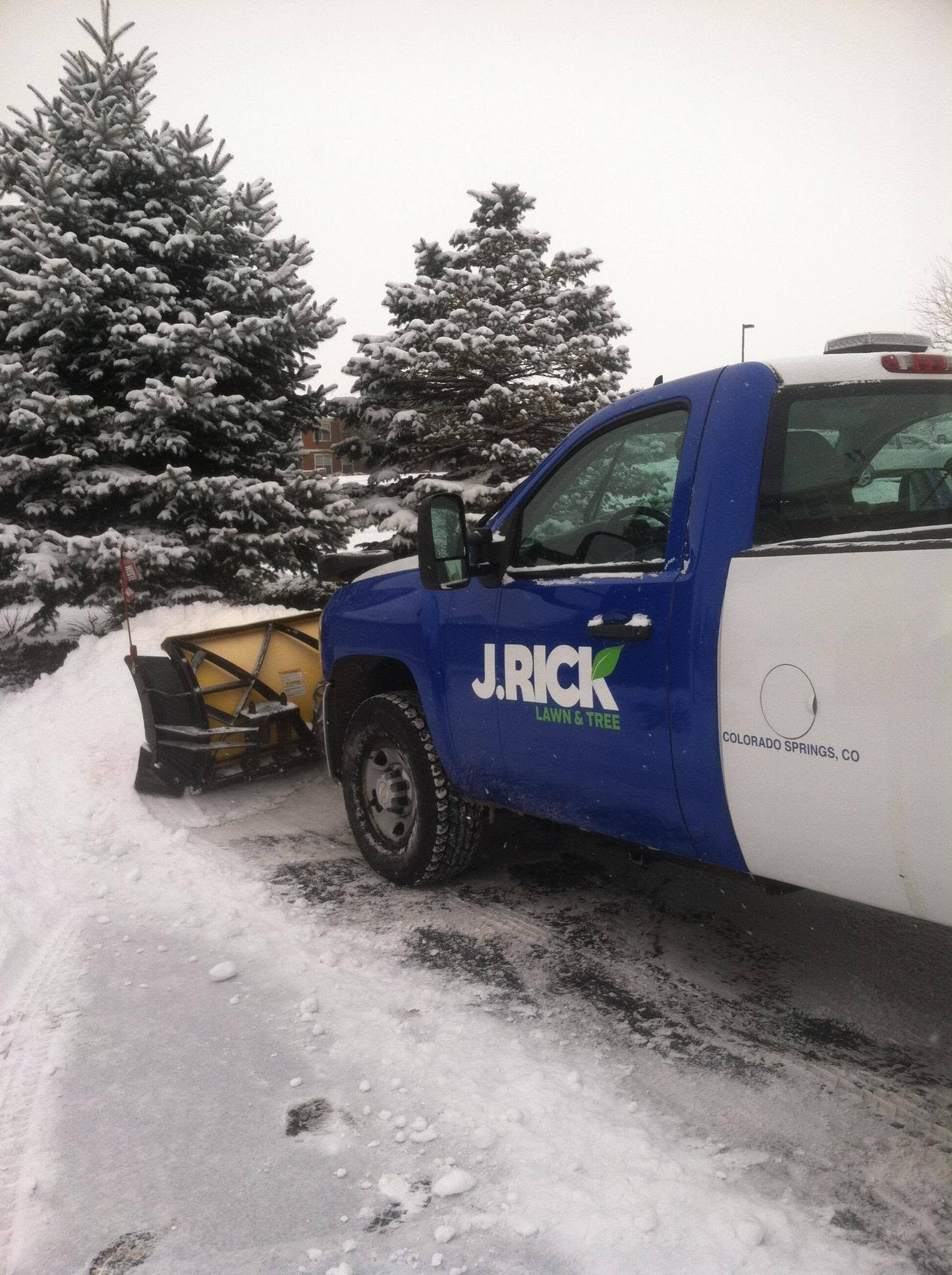 Blue and white JRICK truck with snowplow clearing a snowy driveway beside evergreen trees