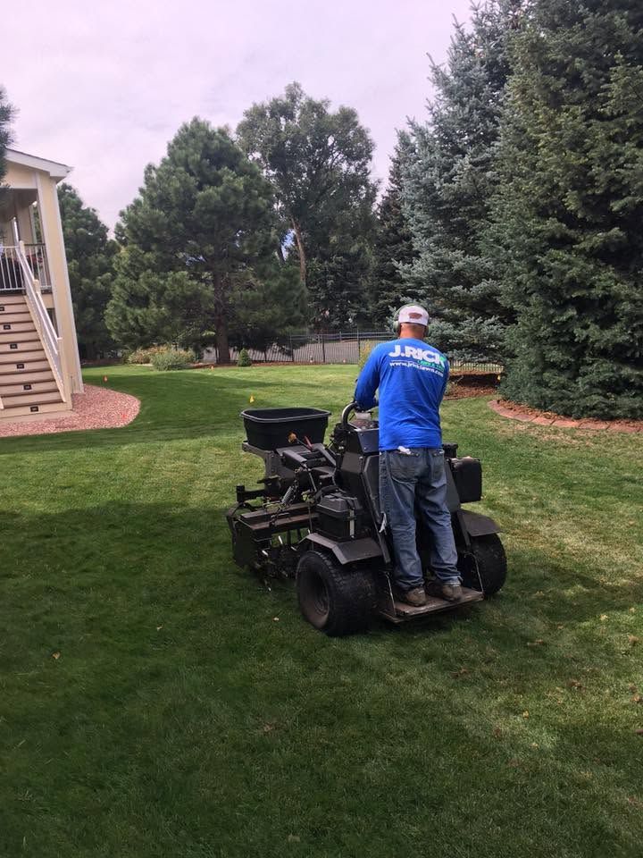 Person in blue shirt on riding mower in a grassy yard near trees and a house staircase