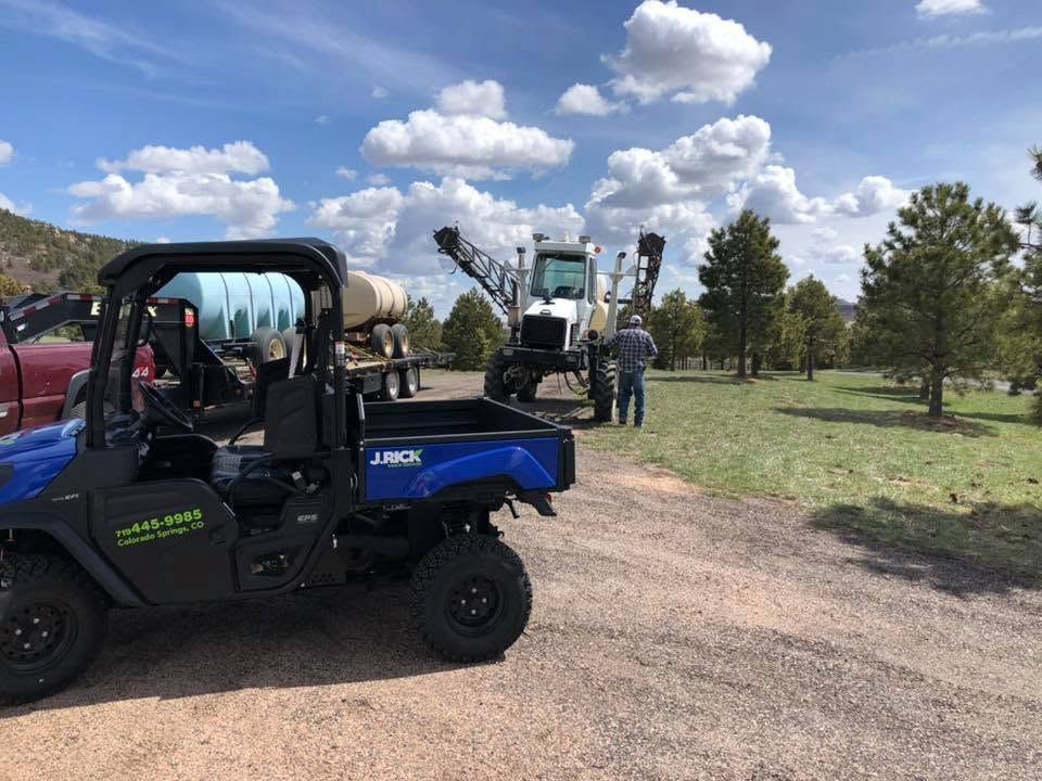 Utility vehicle and people near a grain auger on a rural farm under a cloudy blue sky.