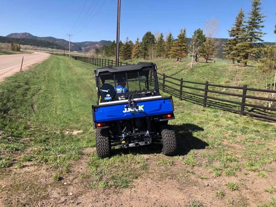 Blue off-road utility vehicle parked on a grassy dirt path beside a fenced field and road, with mountains in the distance