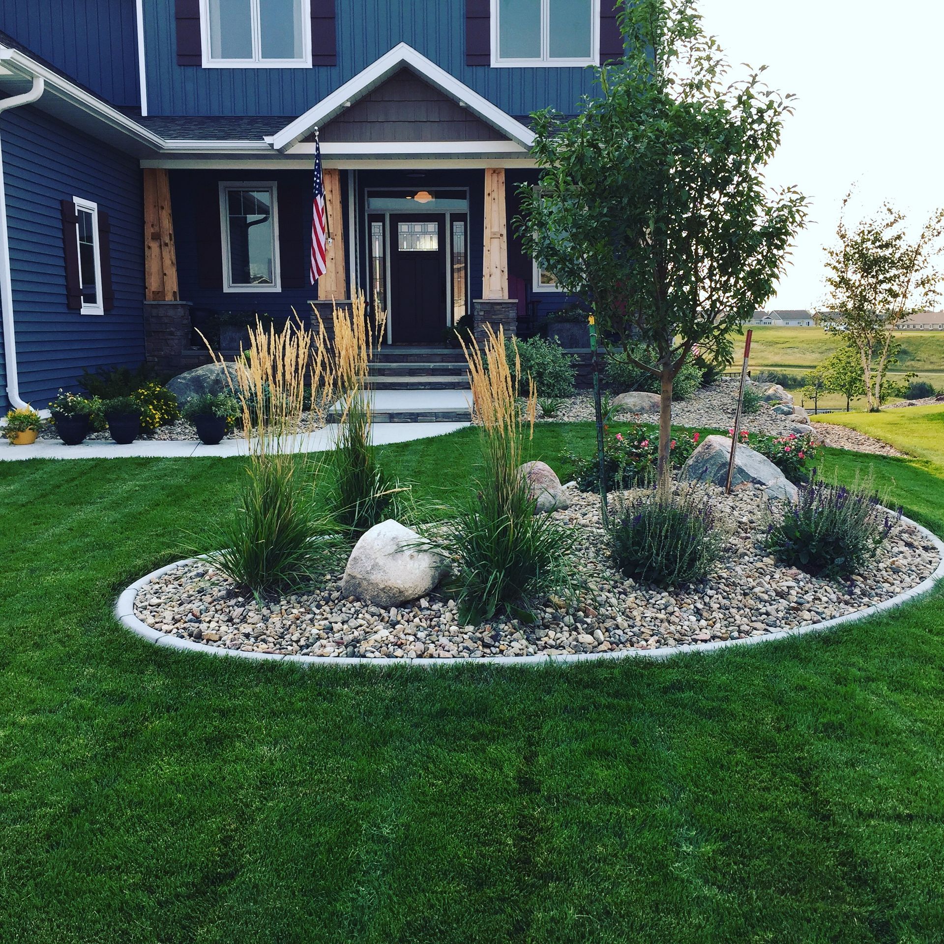 Front yard garden with rocks and grasses in front of a dark blue house with a porch