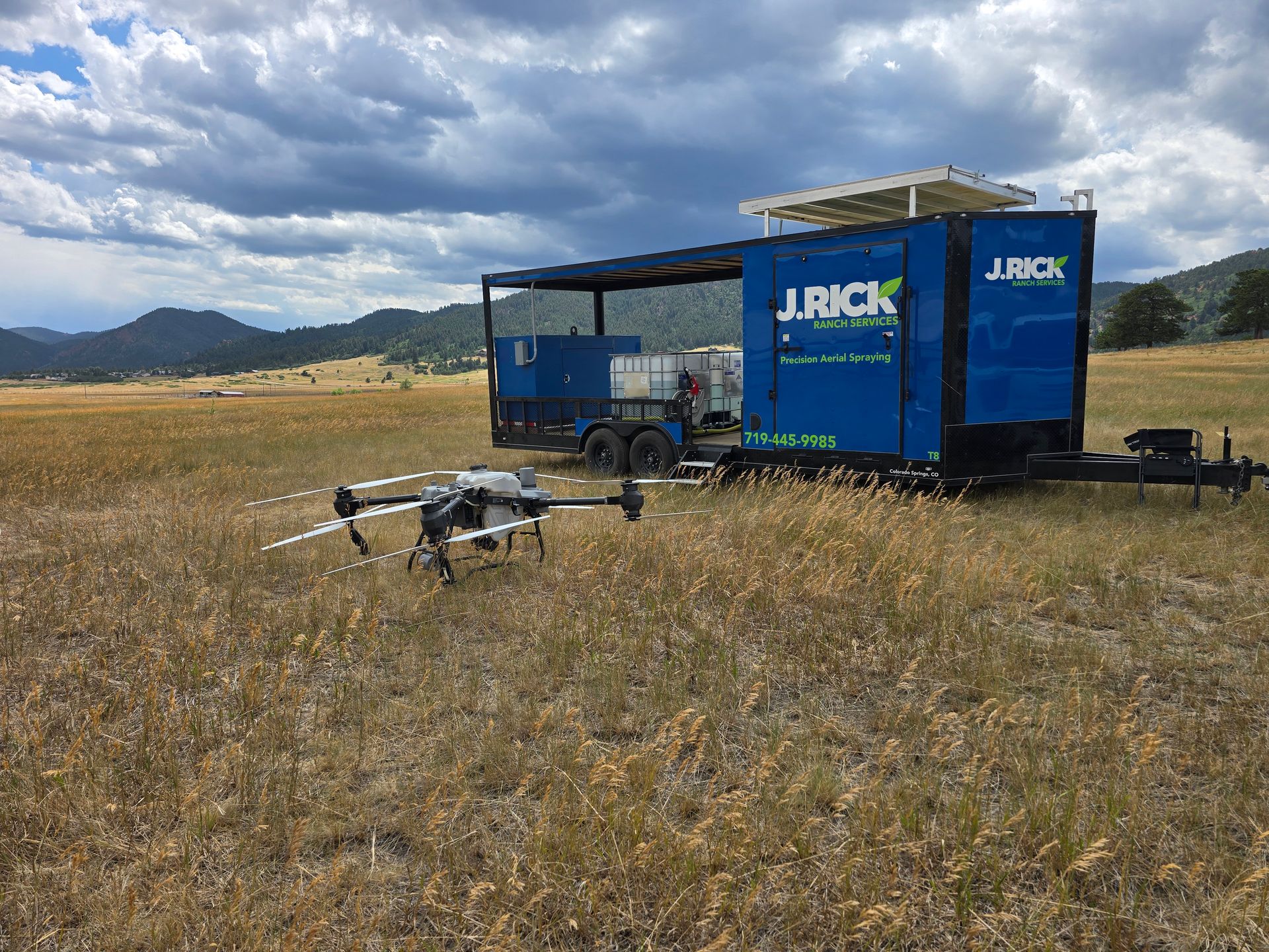 Blue JRICK trailer and equipment on a grassy field under cloudy mountains.