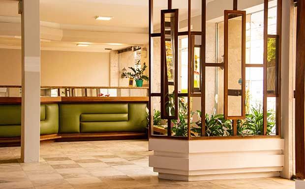 Hall de entrada com assentos verdes, divisória de madeira com plantas e piso de mármore.