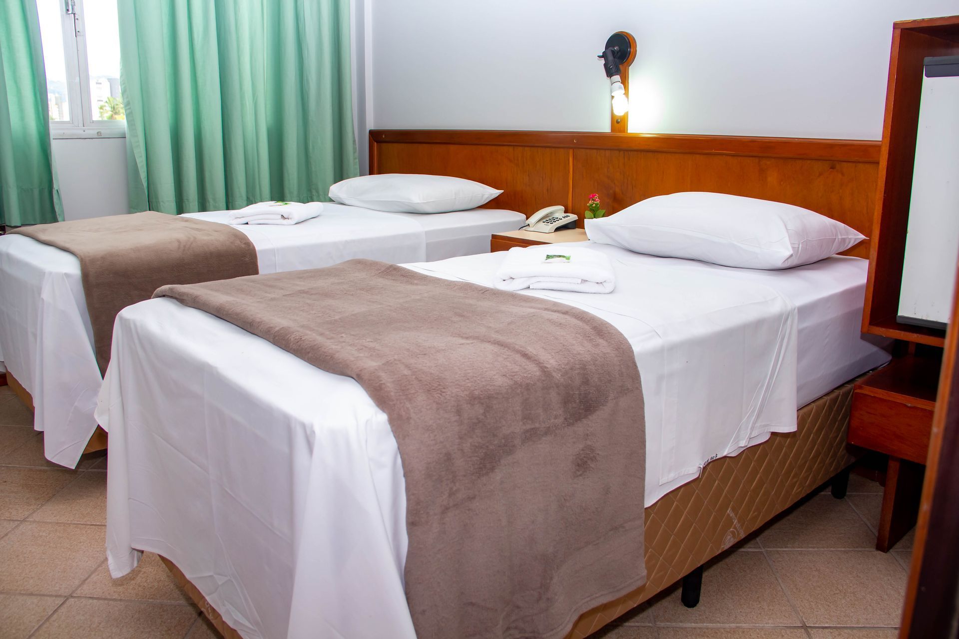 Duas camas de solteiro em um quarto de hotel, com lençóis brancos e cobertores marrons. Cortinas verde-claras.