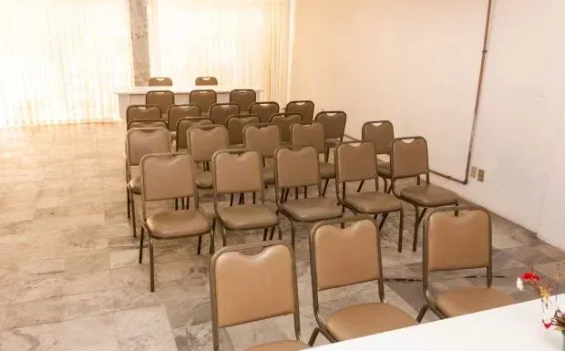 Fileiras de cadeiras bege vazias estão dispostas de frente para uma mesa em uma sala bem iluminada.