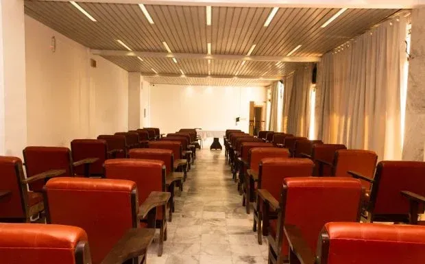 Fileiras de cadeiras vermelhas estão voltadas para uma tela em branco em um auditório iluminado com paredes brancas e piso claro.