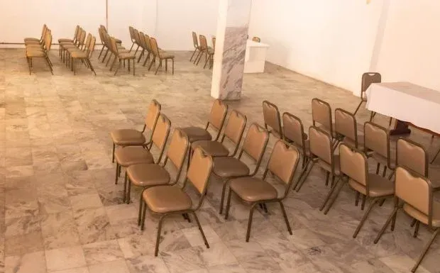 Fileiras de cadeiras vazias em tom bege dispostas em uma sala ampla com piso de mármore e paredes brancas.
