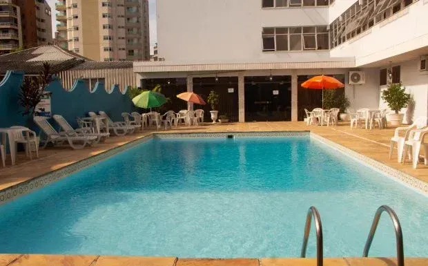 Piscina com espreguiçadeiras, guarda-sóis e prédio ao fundo.