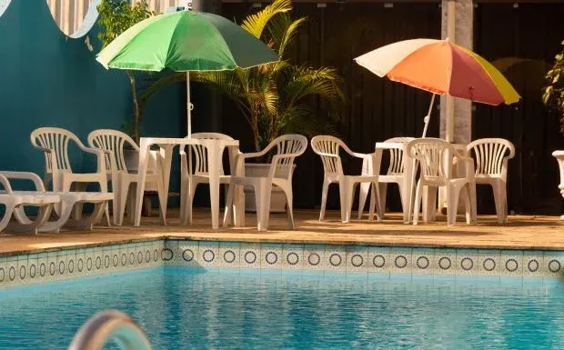 Cena à beira da piscina com mesas e cadeiras de plástico branco sob guarda-sóis verdes e laranjas.