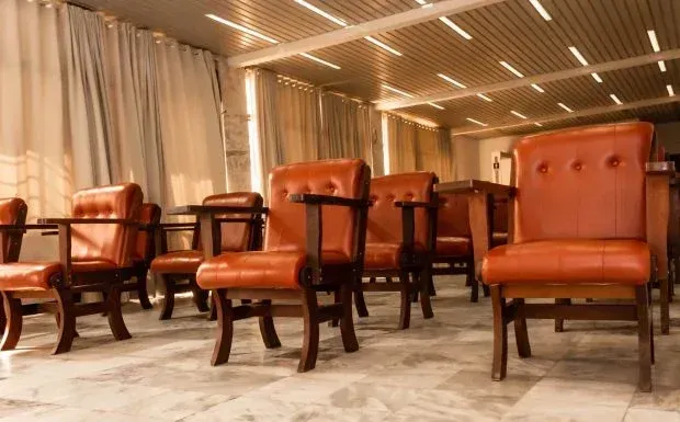 Fileiras de poltronas de couro marrom em uma sala vazia, sob um teto claro.