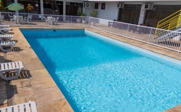 Piscina com água azul rodeada por pedra bege e cadeiras brancas, cerca e um edifício.