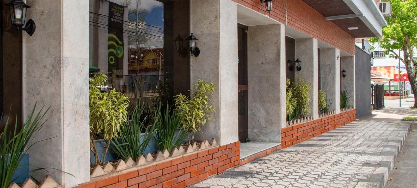 Exterior de um edifício com fachada de tijolos, colunas, janelas e vasos de plantas. Calçada pavimentada.