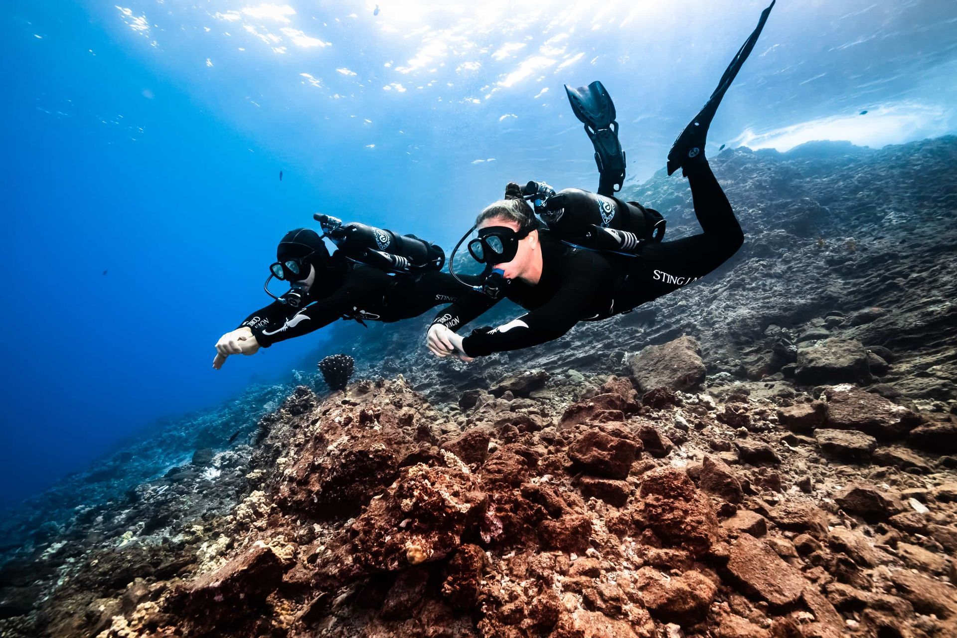 Two Dive Friends scuba divers swim over a rocky coral reef in clear blue water