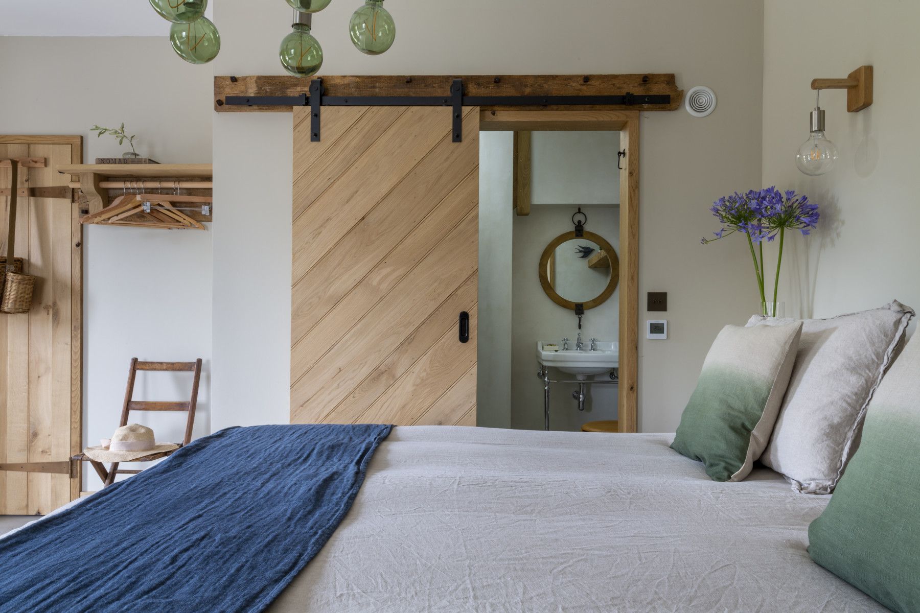 Bedroom with barn door leading to a bathroom. Bed has blue blanket and green-ombre pillows. Light fixtures above.