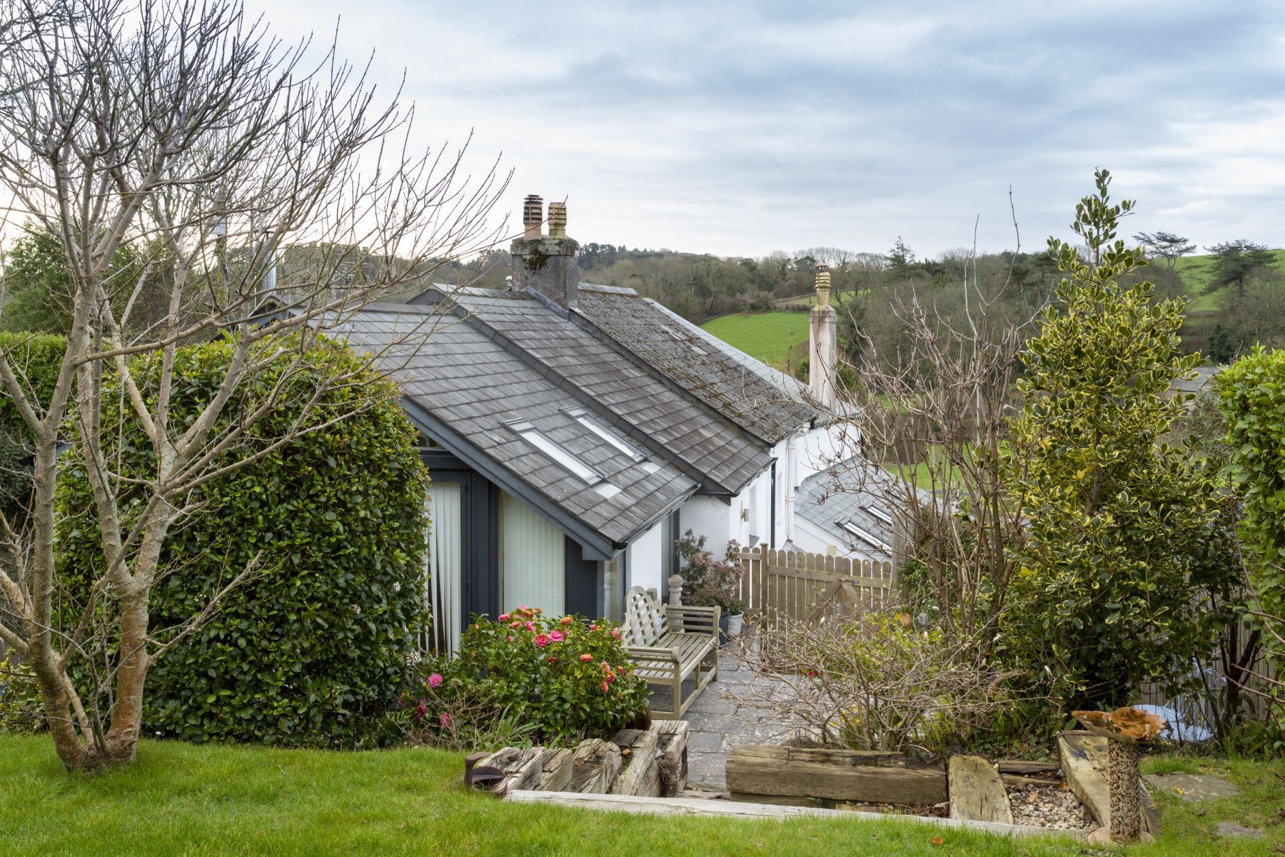 Cottage with a slate roof, set amidst green bushes and trees, with a stone path in the front.