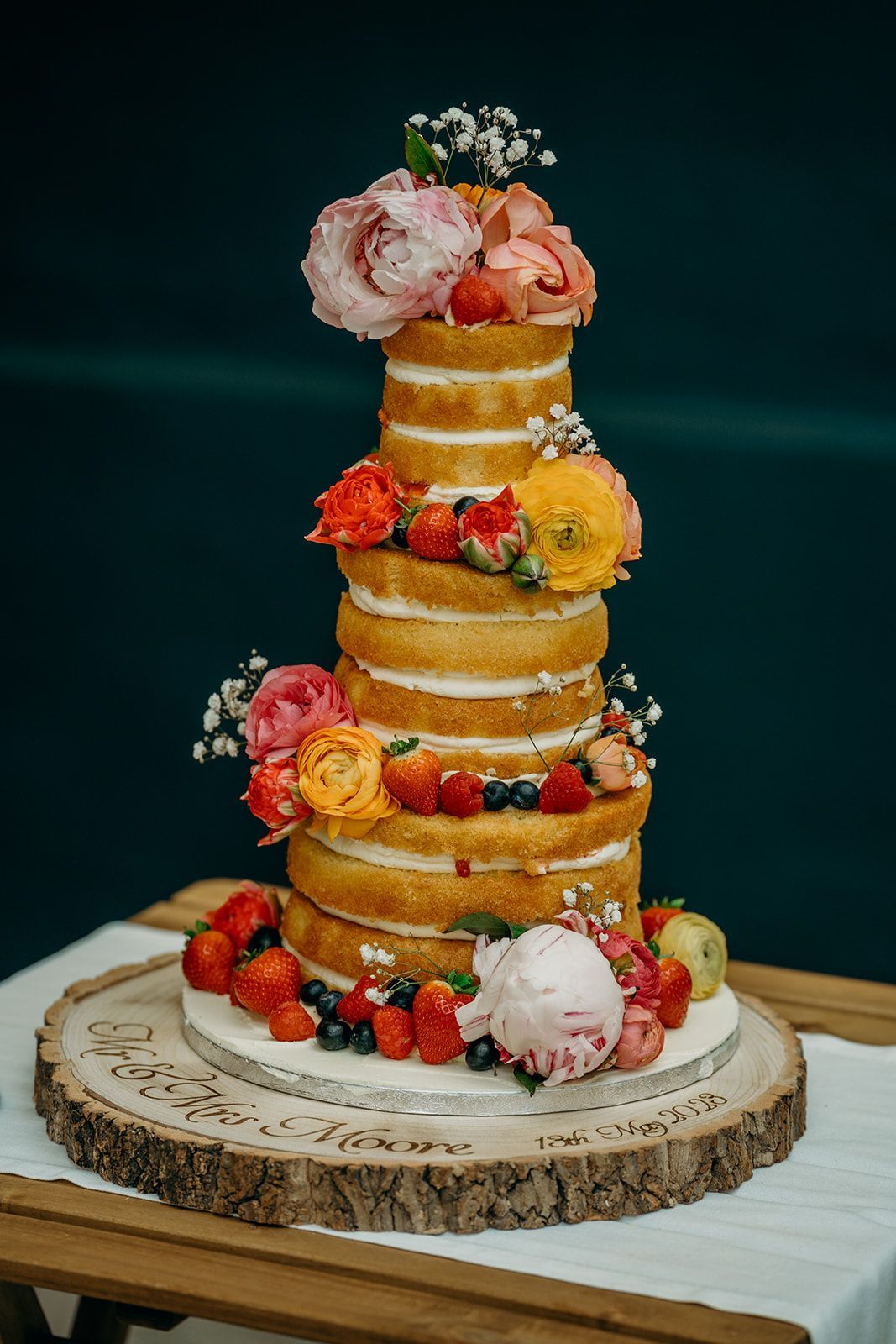 Naked tiered cake with flowers, berries, and a scoop of pink ice cream on a wooden base.