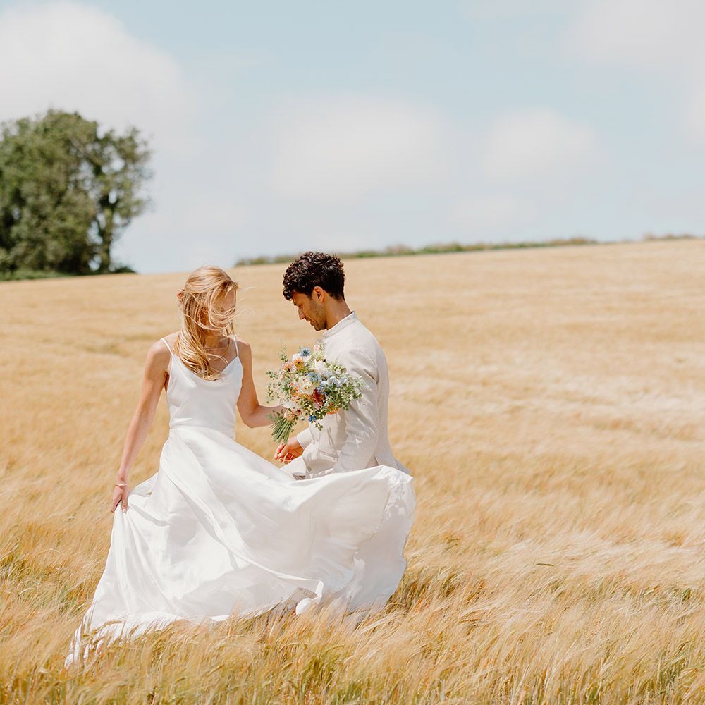 Couple walking in a golden field, the bride in a white dress carrying a bouquet, on a sunny day.