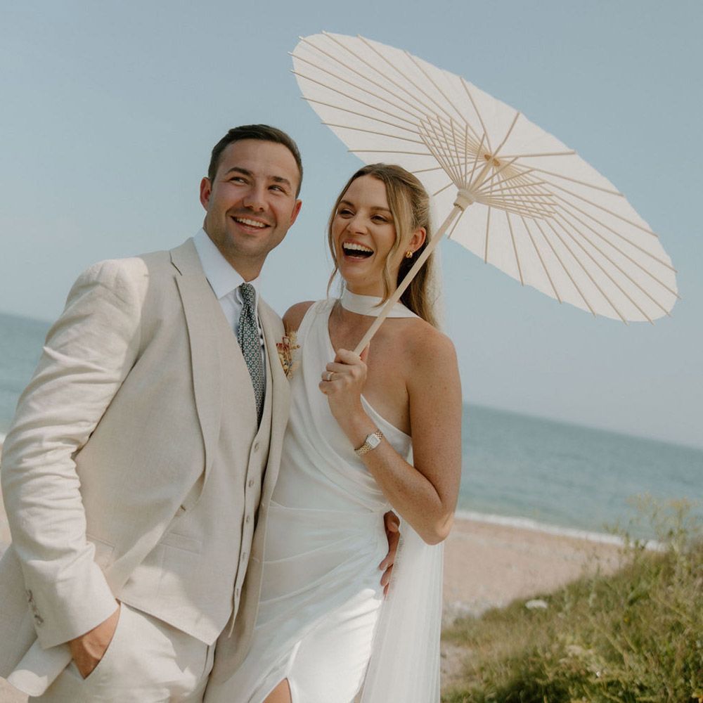 Wedding couple smiling near the ocean, woman holding white parasol, man in linen suit.