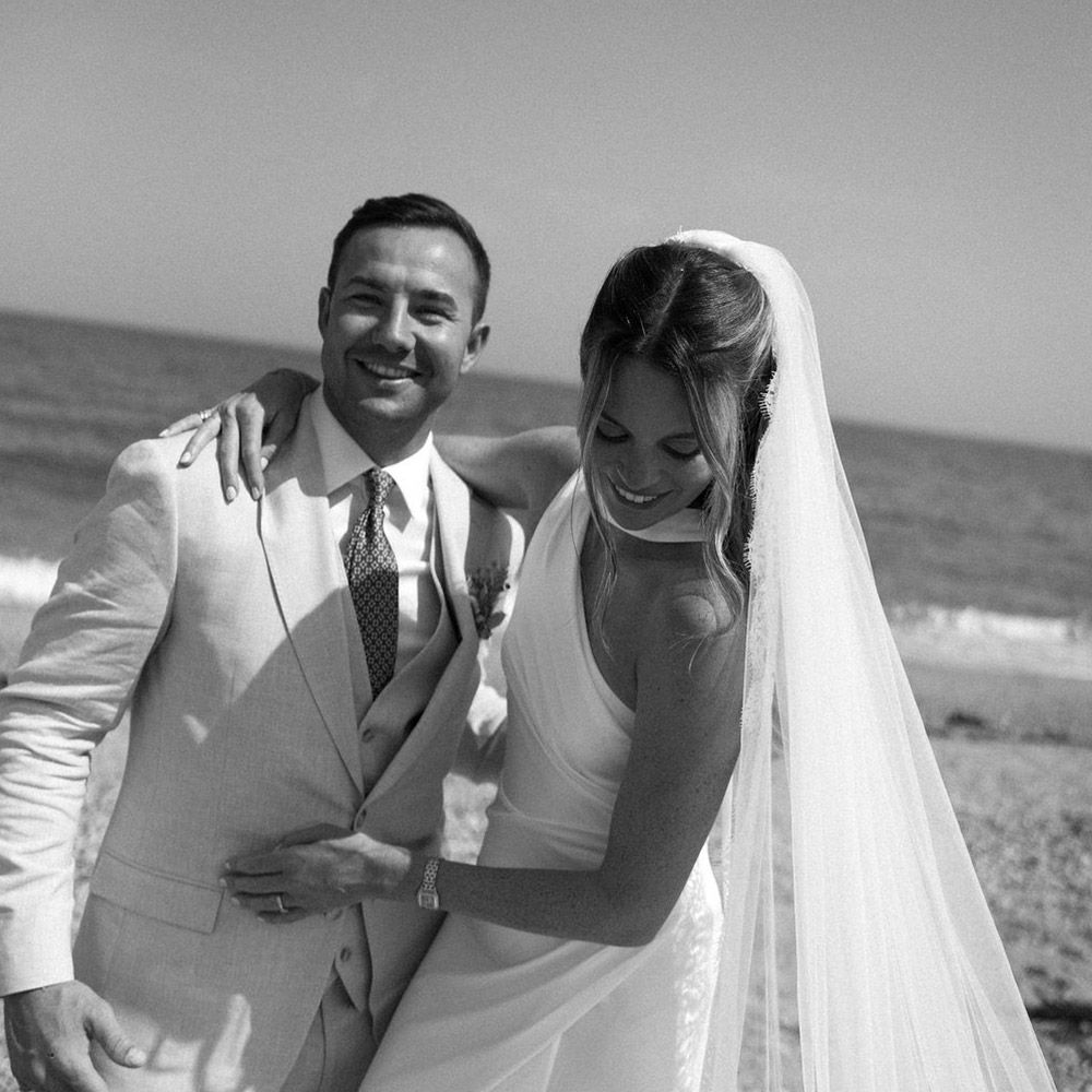 Bride and groom pose on beach, bride in white dress and veil, groom in tan suit.