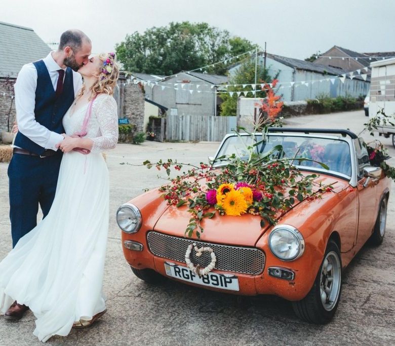 Wedding couple kissing by an orange convertible decorated with flowers in a rural setting.