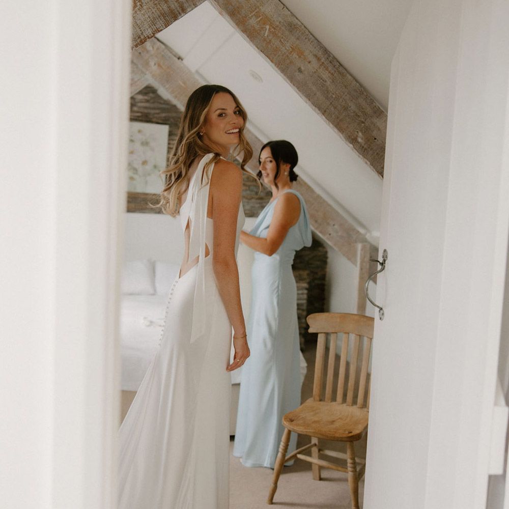 Bride in white dress smiles while bridesmaid adjusts dress in a bright room with wood beams.