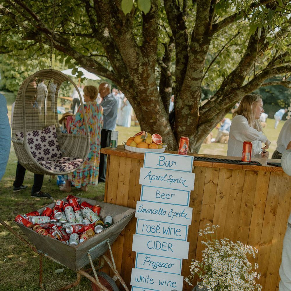 Outdoor bar with drinks menu. Wheelbarrow of cans, people socializing near tree.