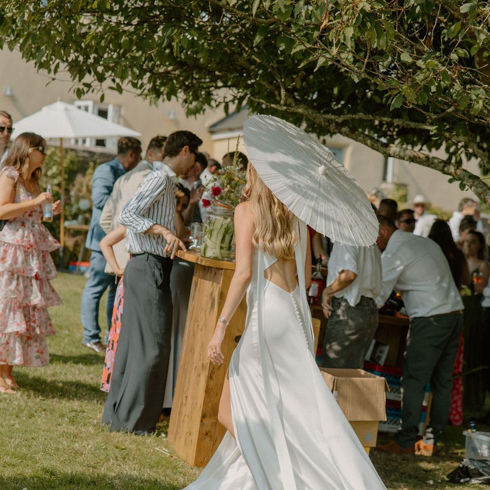 Woman in white dress and parasol at outdoor event, near a wooden bar. People in background.