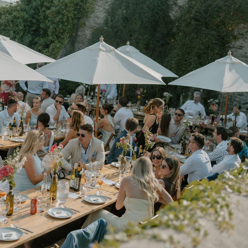 Outdoor wedding reception with guests seated at long tables under umbrellas.