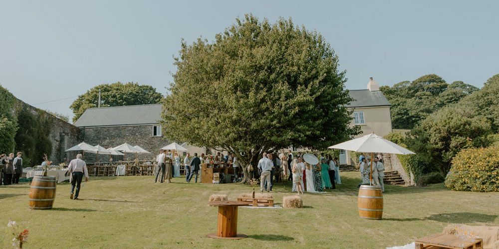 Wedding guests gather outdoors; tables with umbrellas, a tree, and a building are visible.