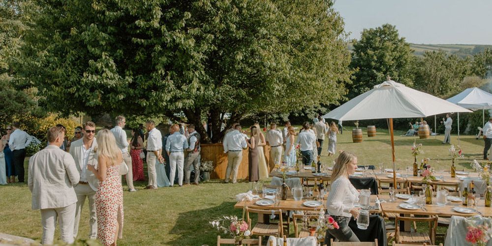 Wedding reception on a grassy lawn with guests, tables, umbrellas, and a large tree.
