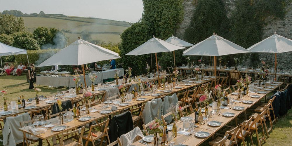 Long tables set for outdoor dining under umbrellas on a grassy lawn.